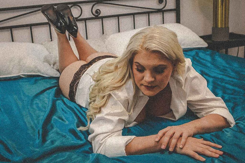 Person with long blonde hair in a white shirt and patterned lingerie, lying on a bed with blue sheets, facing the camera.