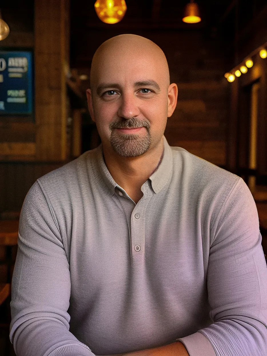 A man with a shaved head and goatee is sitting at a table in a warmly lit room, wearing a light gray collared sweater.